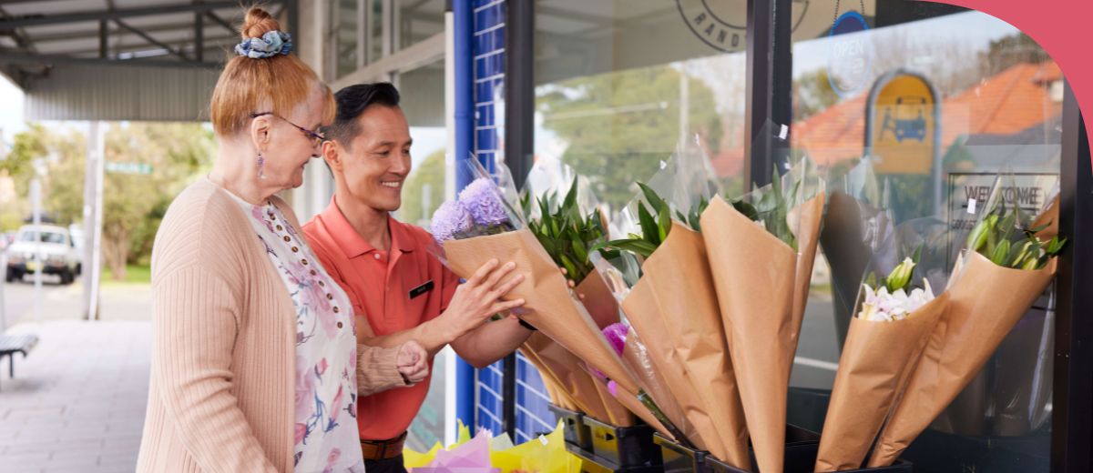 An older woman and younger man stand together in a street in front of a shop with a display of flower bouquets. They both look at a bouquet that the man is pulling out of the display.