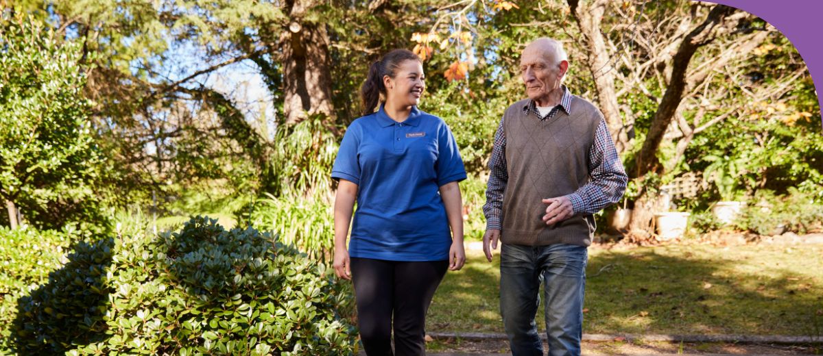 An older man and younger woman walk side by side in a garden, smiling.
