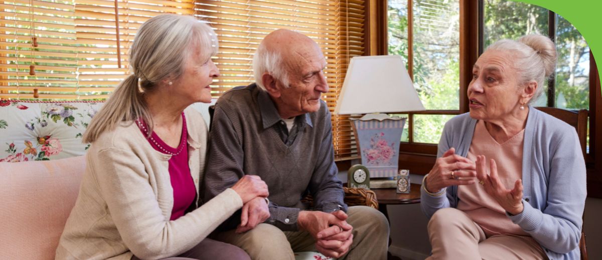 3 older people sit together in a lounge room, talking.
