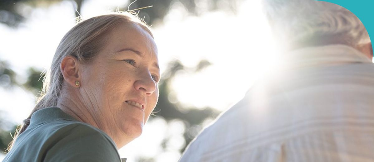 View from behind 2 people. A woman on the left looks at a person on the right. All that can be seen of the other person is short grey hair.