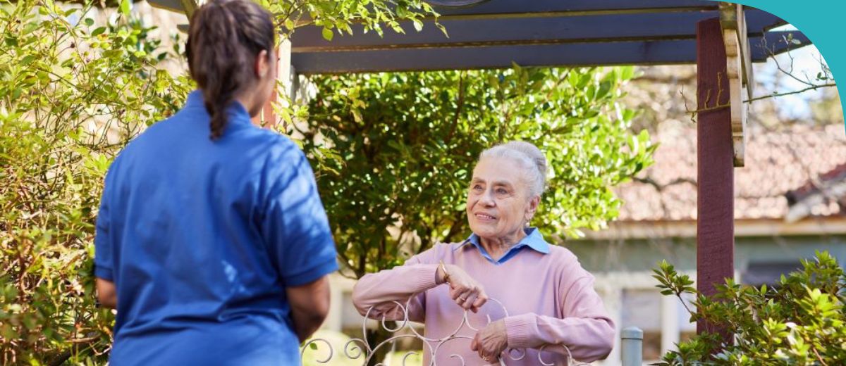 An older woman with white hair, standing by a gate with the sign "Rose Cottage". She is wearing a light purple sweater over a blue collared shirt. Facing her is a caregiver in a blue uniform with her back to the camera. The setting appears to be a sunny residential garden, and it seems they are engaged in a conversation or an outdoor activity.