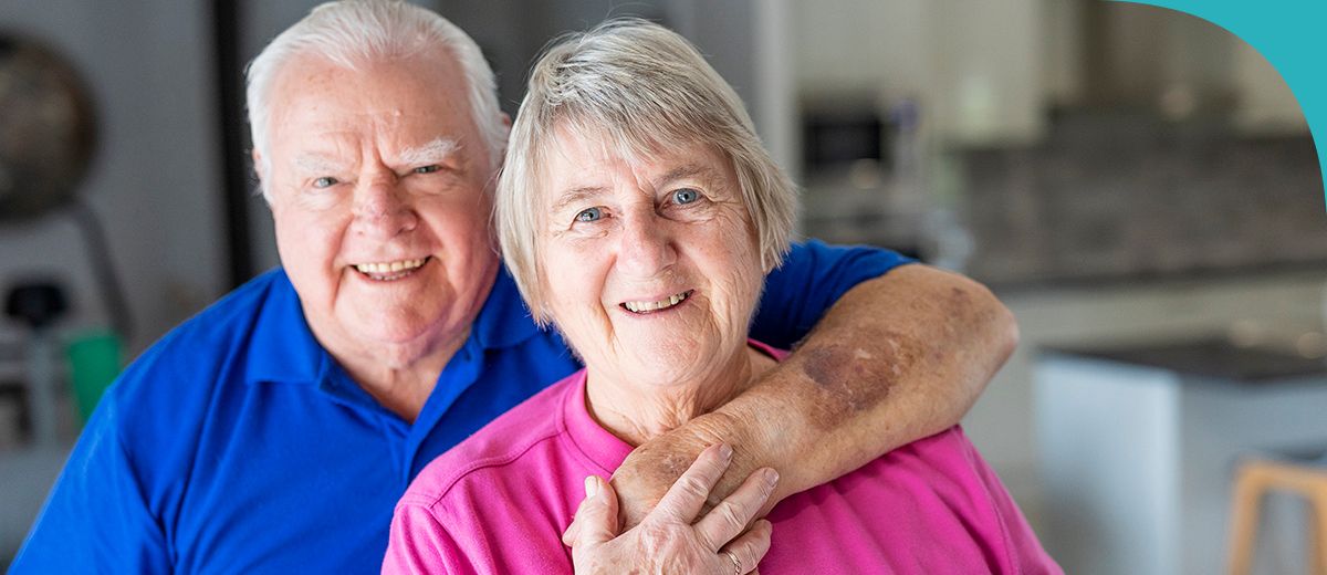 An older couple smiles warmly at the camera, with the man wearing a blue polo shirt and the woman in a pink shirt. The man has his arm around the womans shoulders, showing a loving and supportive gesture.
