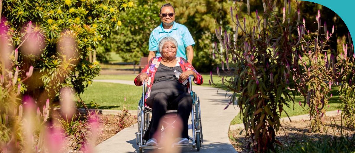 A smiling older woman is being pushed in a wheelchair by a caregiver along a garden pathway. The scene is vibrant with greenery and pink flowers on both sides, creating a peaceful and joyful atmosphere.