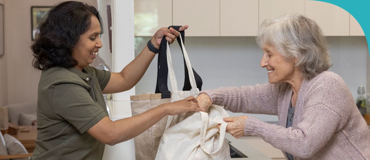 2 women stand on opposite sides of a kitchen bench. One woman is placing canvas grocery bags on the bench and the other, who is older, is opening a bag and reaching her hand in.