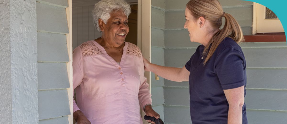An older woman stands in a doorway with a walking frame. A younger woman stands in front of her with a hand on her arm and carrying a bad in her other hand. They are looking at each other and smiling.