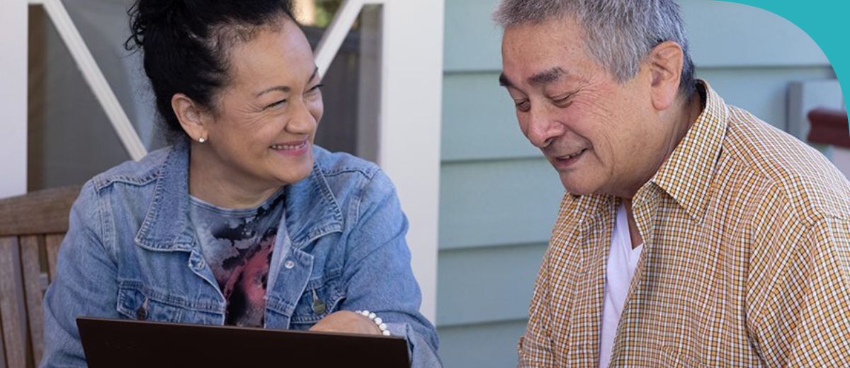 A man and a woman, both senior, sit outside in front of a laptop, both smiling.