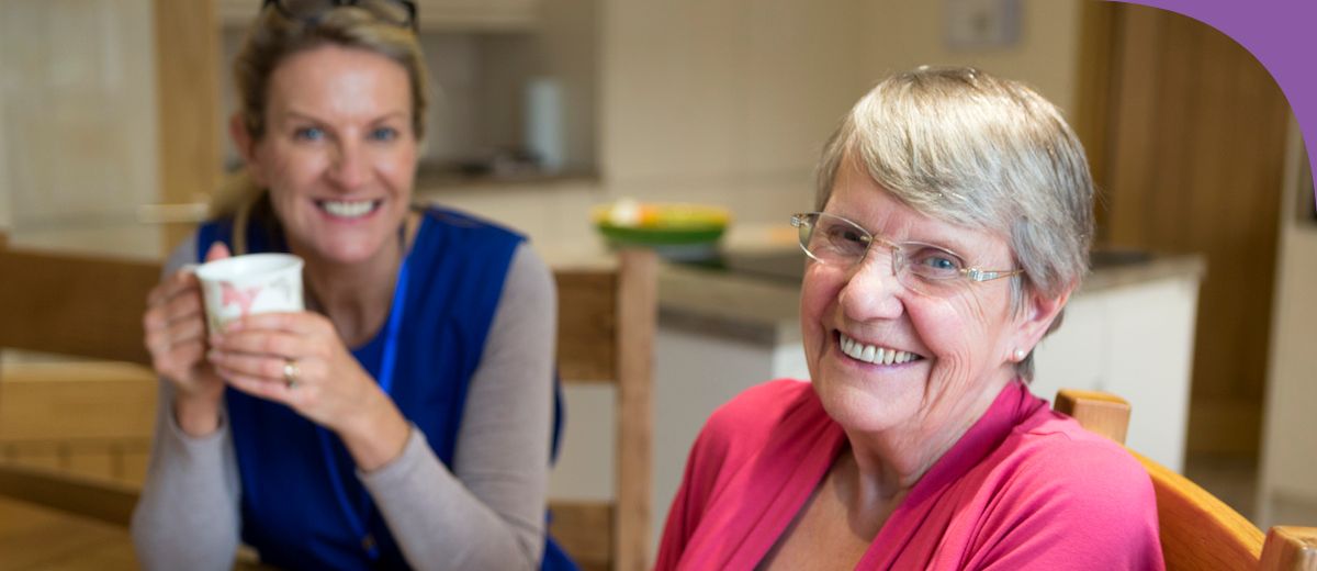 Two women smiling and enjoying a conversation over cups of tea in a well-lit kitchen.