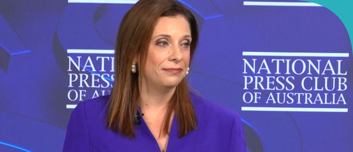 A lady speaking in front of a purple banner that reads National Press Club