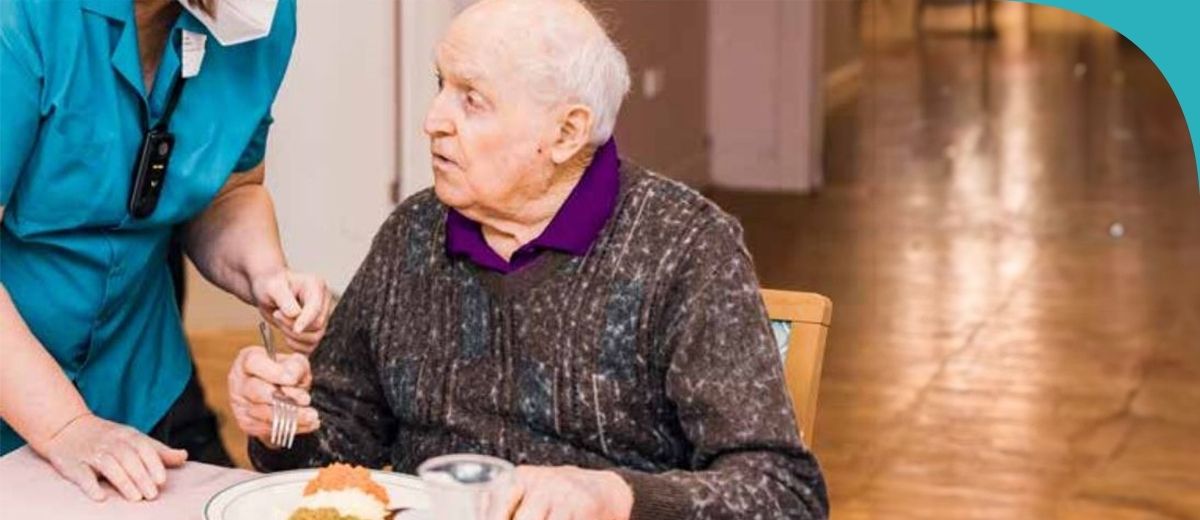 An older man having a meal. An aged care worker is with him. and talking