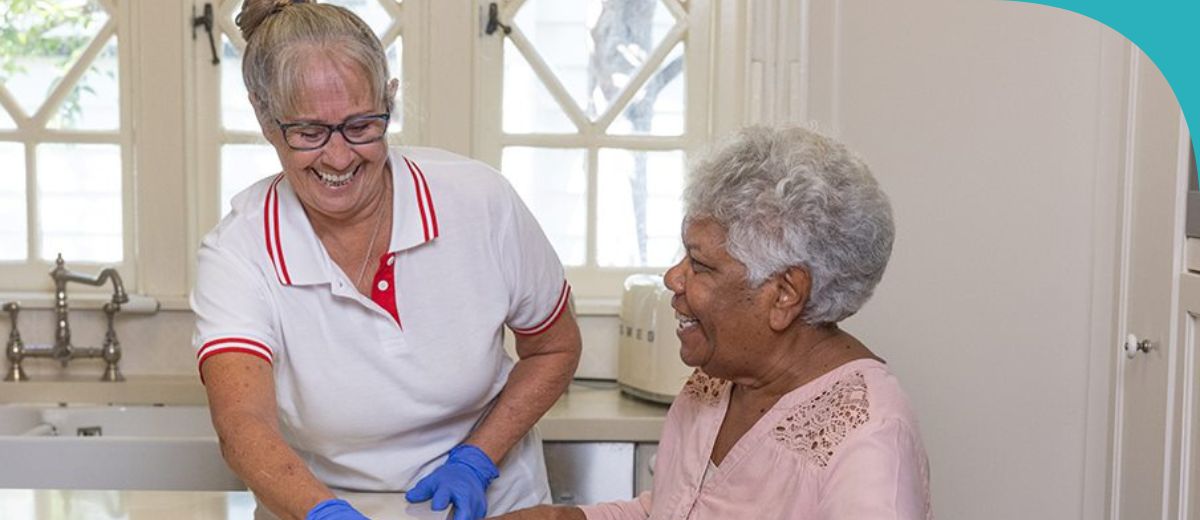 An older woman sits in a walking frame at a kitchen bench. Another woman stands next to her at the bench, wiping it with a cloth. Both are smiling.