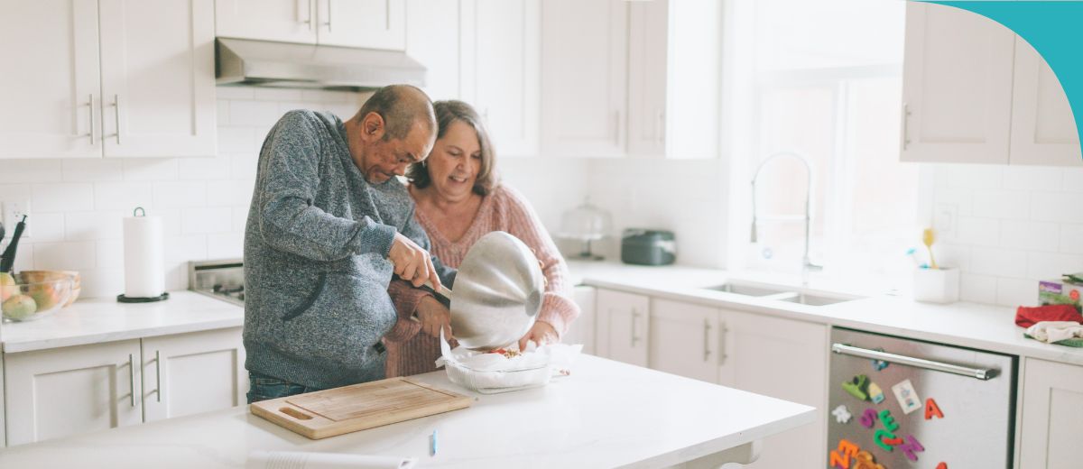 A man and a woman, both seniors, standing together in a kitchen. The woman holds a bowl on the kitchen counter while the man scrapes something into it from another bowl.