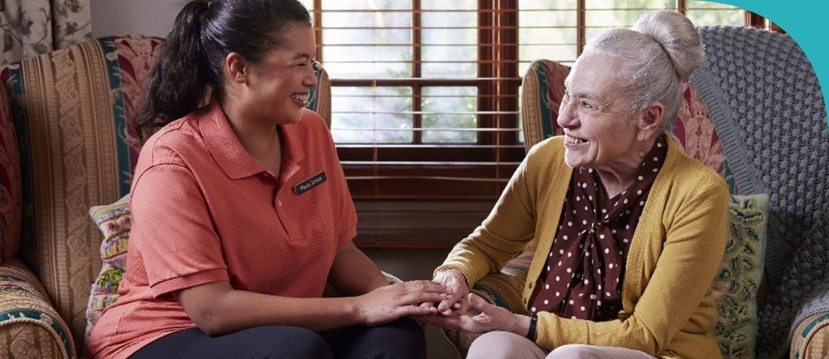 A cheerful young woman in a coral-coloured polo shirt with a nametag sits beside an elderly lady in a mustard cardigan and polka-dot blouse. They are holding hands, sharing a genuine moment of connection and joy, while seated on colourful patterned armchairs inside a cozy room.