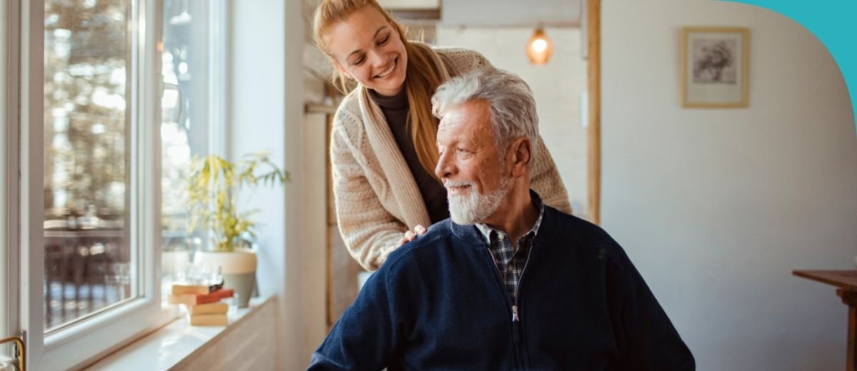 A young woman with blonde hair, wearing a beige knitted cardigan, stands by a window, leaning down to place a comforting hand on the shoulder of an elderly man with grey hair and a navy blue shirt. They both share a moment of warmth and understanding in a well-lit room with a serene outdoor view.