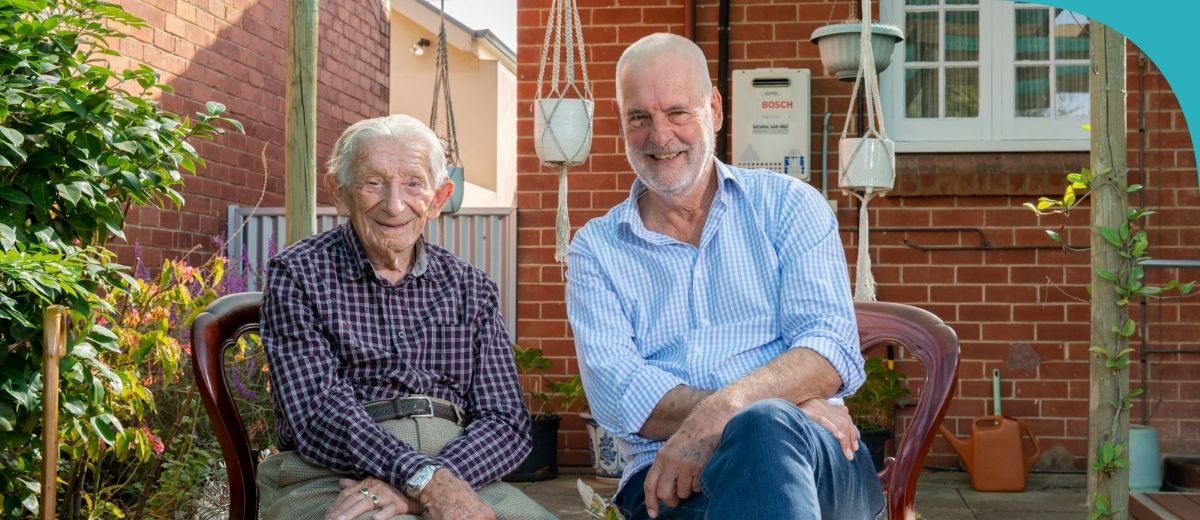 An older man, dressed in a maroon and white checked shirt, sits on a wooden chair in a backyard garden, smiling contentedly. Beside him stands a middle-aged man in a light blue striped shirt, with a relaxed posture and a cheerful expression. They are set against a backdrop of a brick house with a few hanging plants